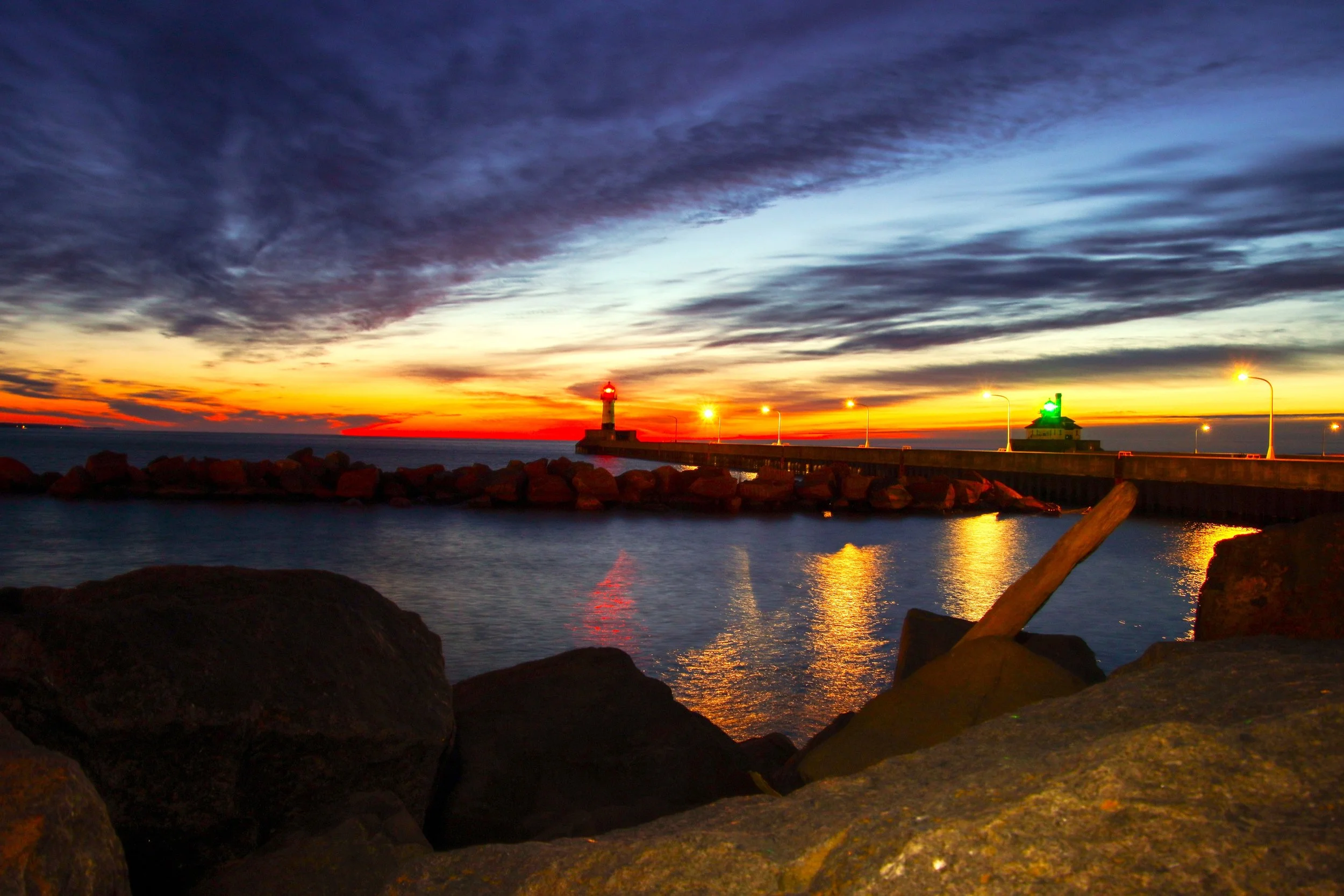 Sunset over the ocean with dark clouds, a lighthouse on a pier, and rocks in the foreground.
