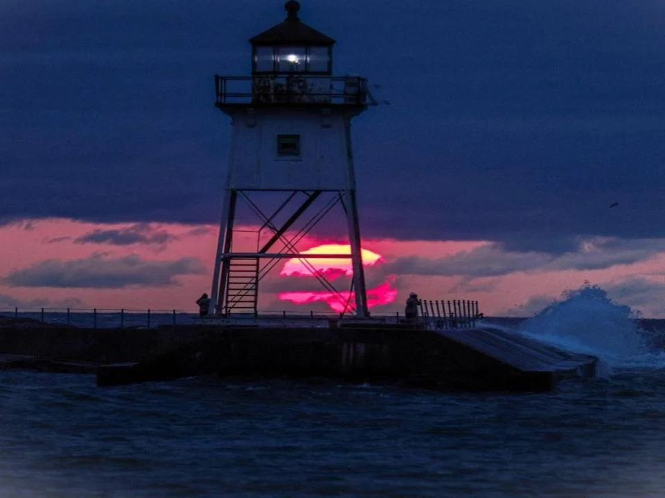Lighthouse at sunset with pink and purple hues, waves crashing on the shore, and two people sitting nearby.
