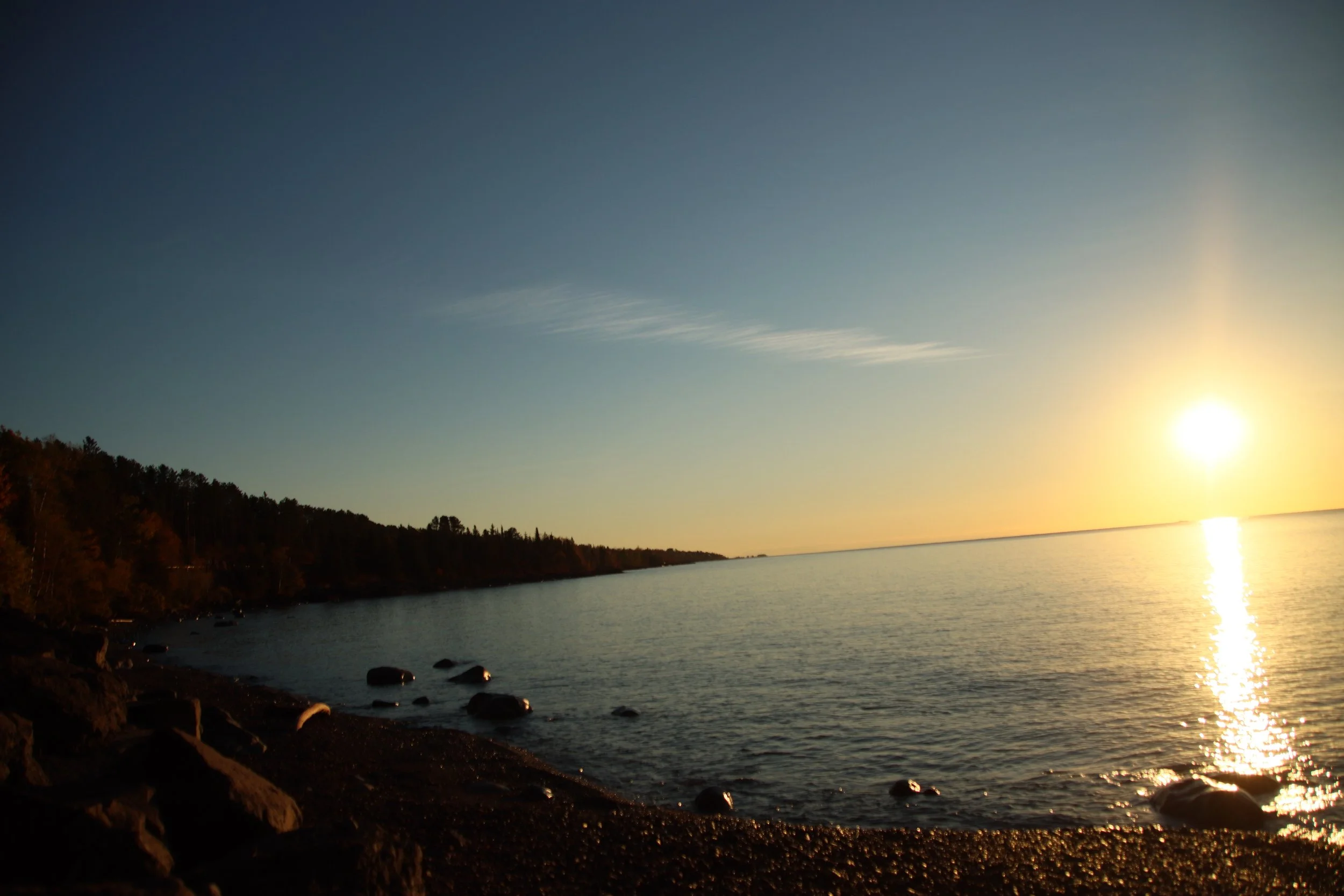 Sunset over a calm body of water with a tree-lined shoreline in the foreground and a mostly clear sky with some wispy clouds.