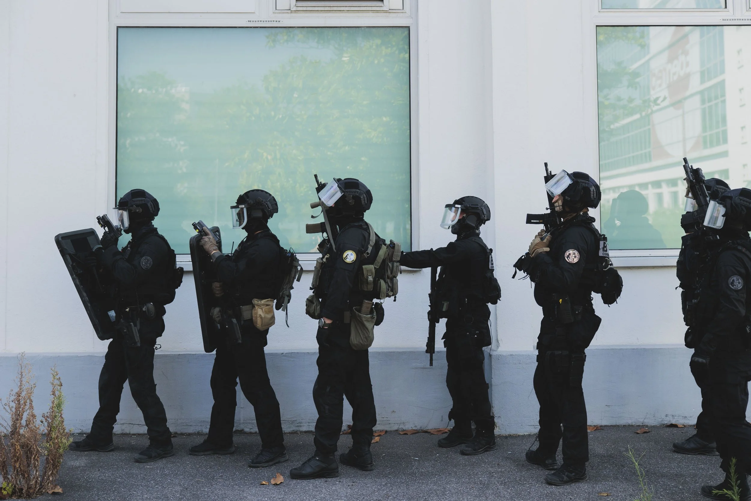 Une ligne de policiers en tenue d'assaut lourd passant devant un bâtiment, portant des casques avec visières et des boucliers, prêts à intervenir.