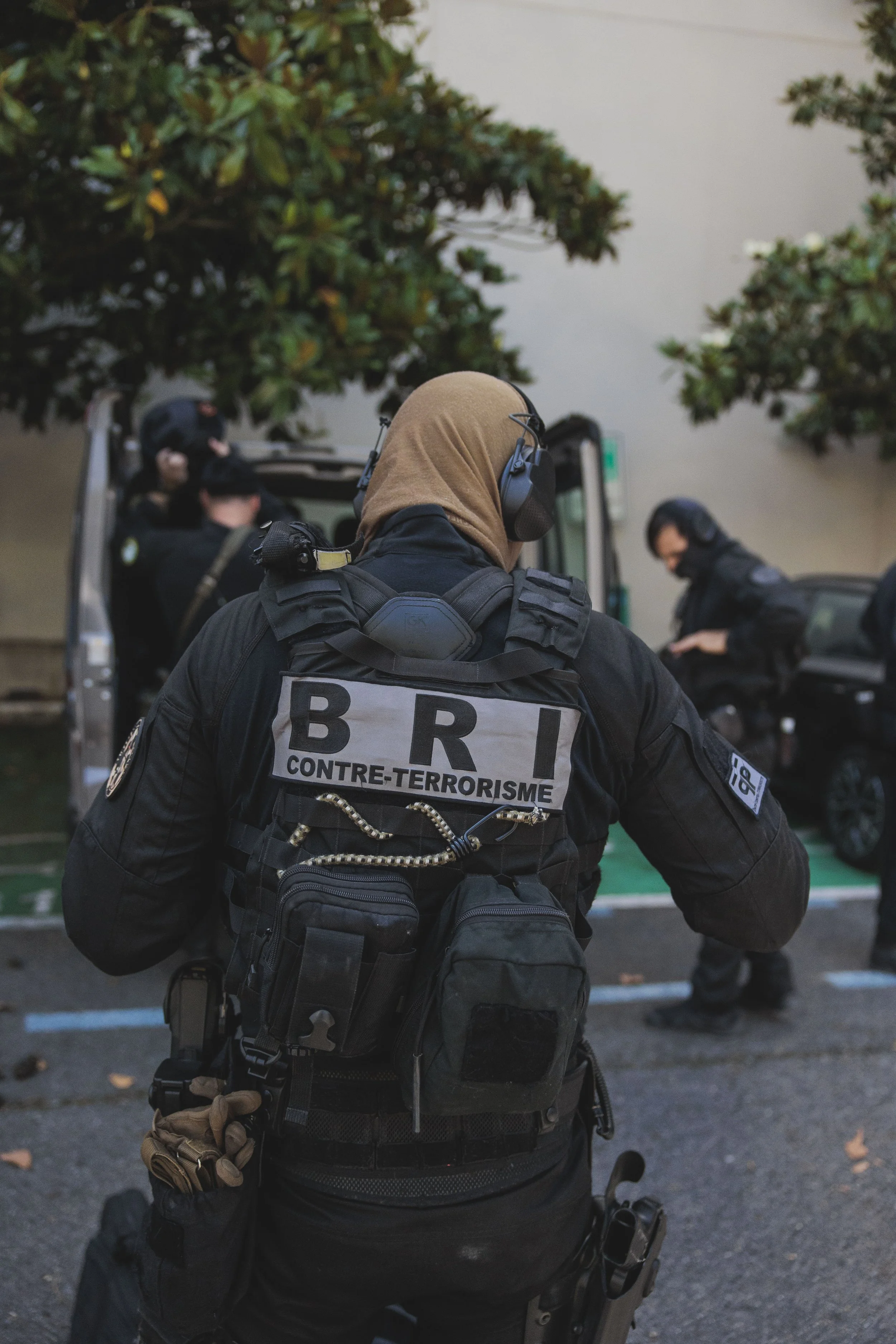 Policier en uniforme paramilitaire portant un gilet pare-balles avec l'inscription Brigade de Recherche et d'Intervention, équipé de matériel tactique, en train de se diriger vers un groupe de policiers devant un véhicule, avec des arbres en fond.