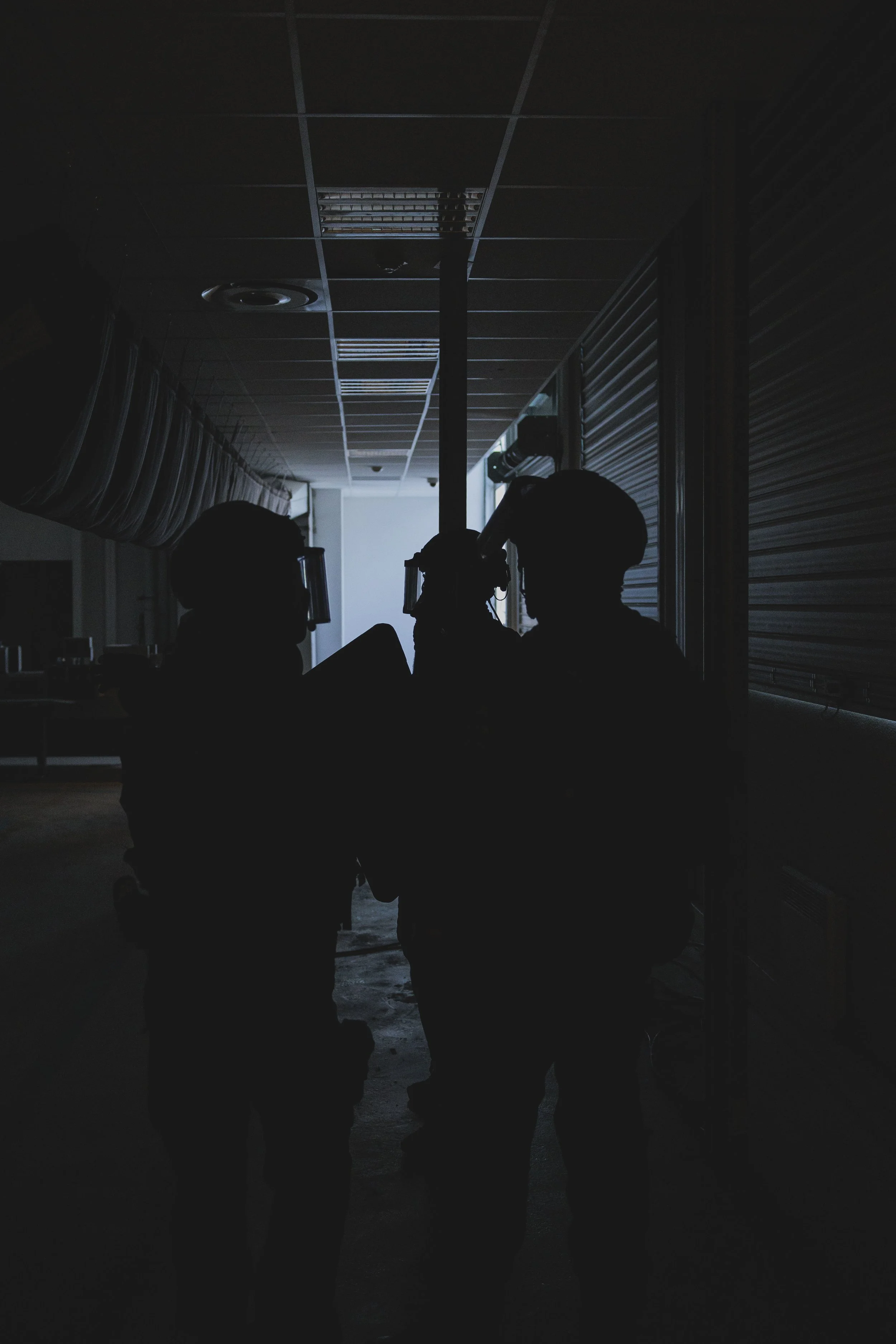 Silhouettes de trois policiers de la BRI-PP en tenue d'assaut lourd dans un couloir sombre, avec un plafond suspendu et des volets métalliques sur le côté.