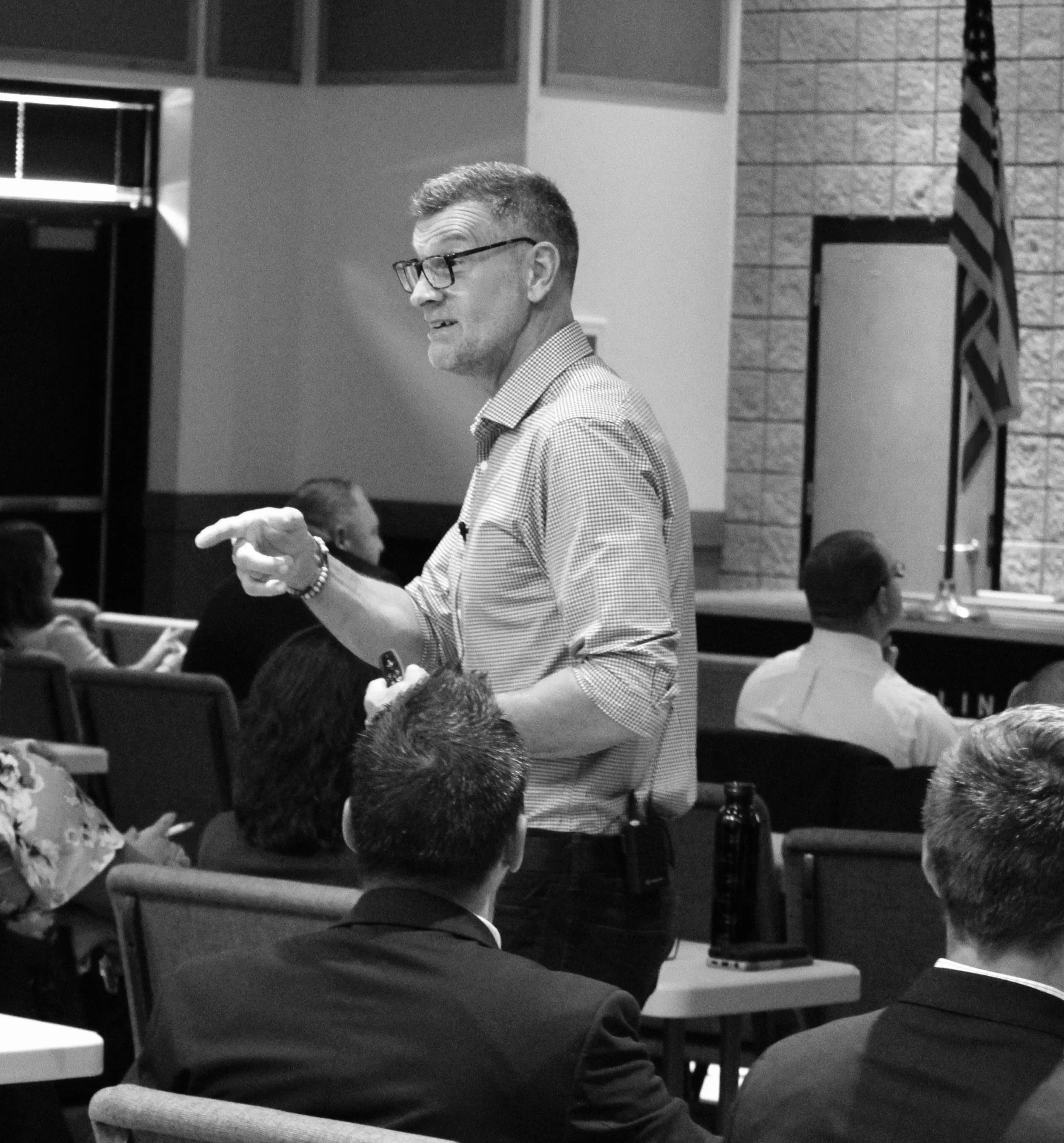 A man with glasses, short hair, wearing a checkered shirt, standing and talking to a group in a room with brick and panel walls, and an American flag.