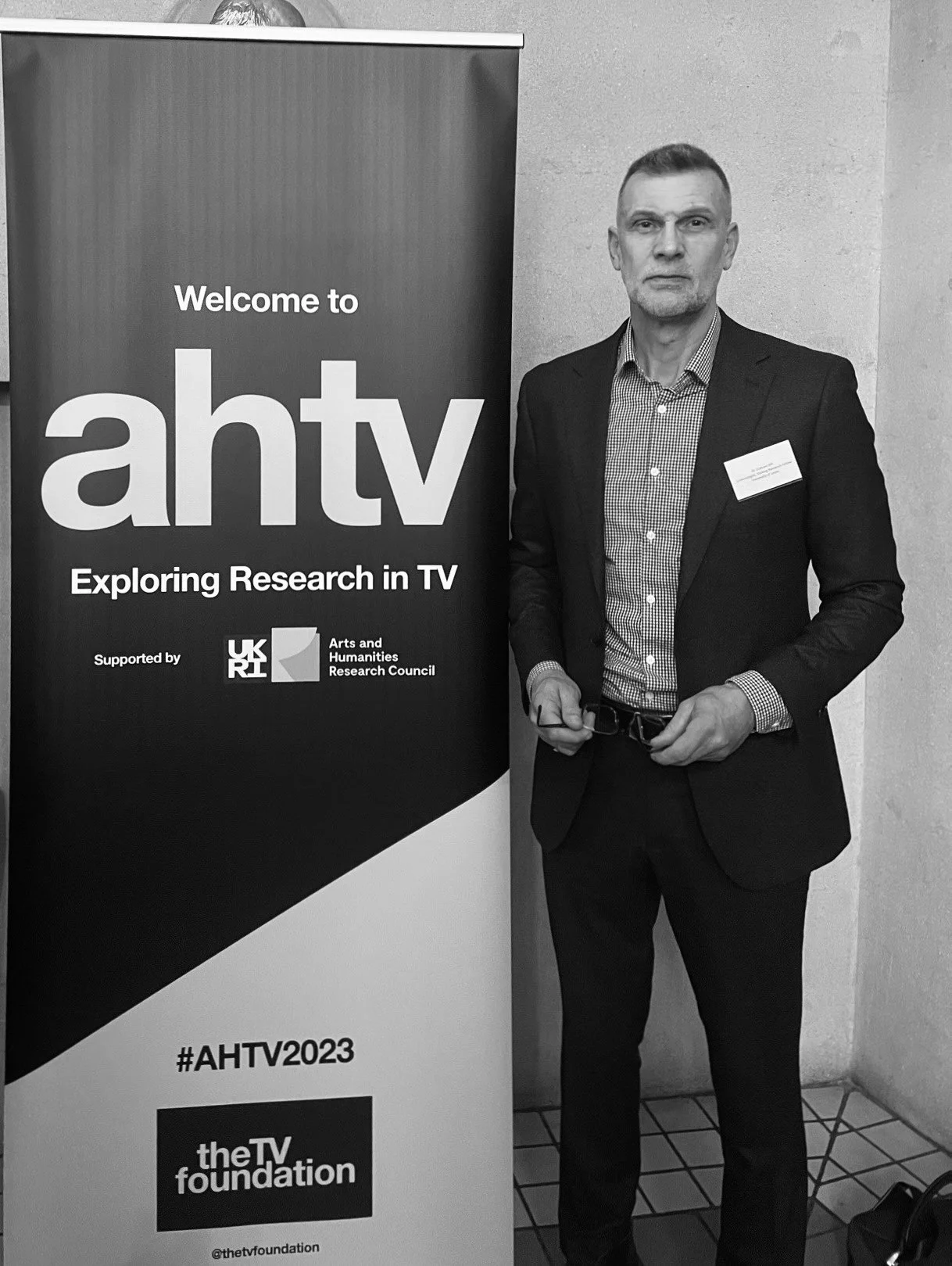 A man in a suit standing next to a banner that reads 'Welcome to AHTV, Exploring Research in TV,' supported by UKRI and the Arts and Humanities Research Council, with the hashtag #AHTV2023 and the TV Foundation logo.