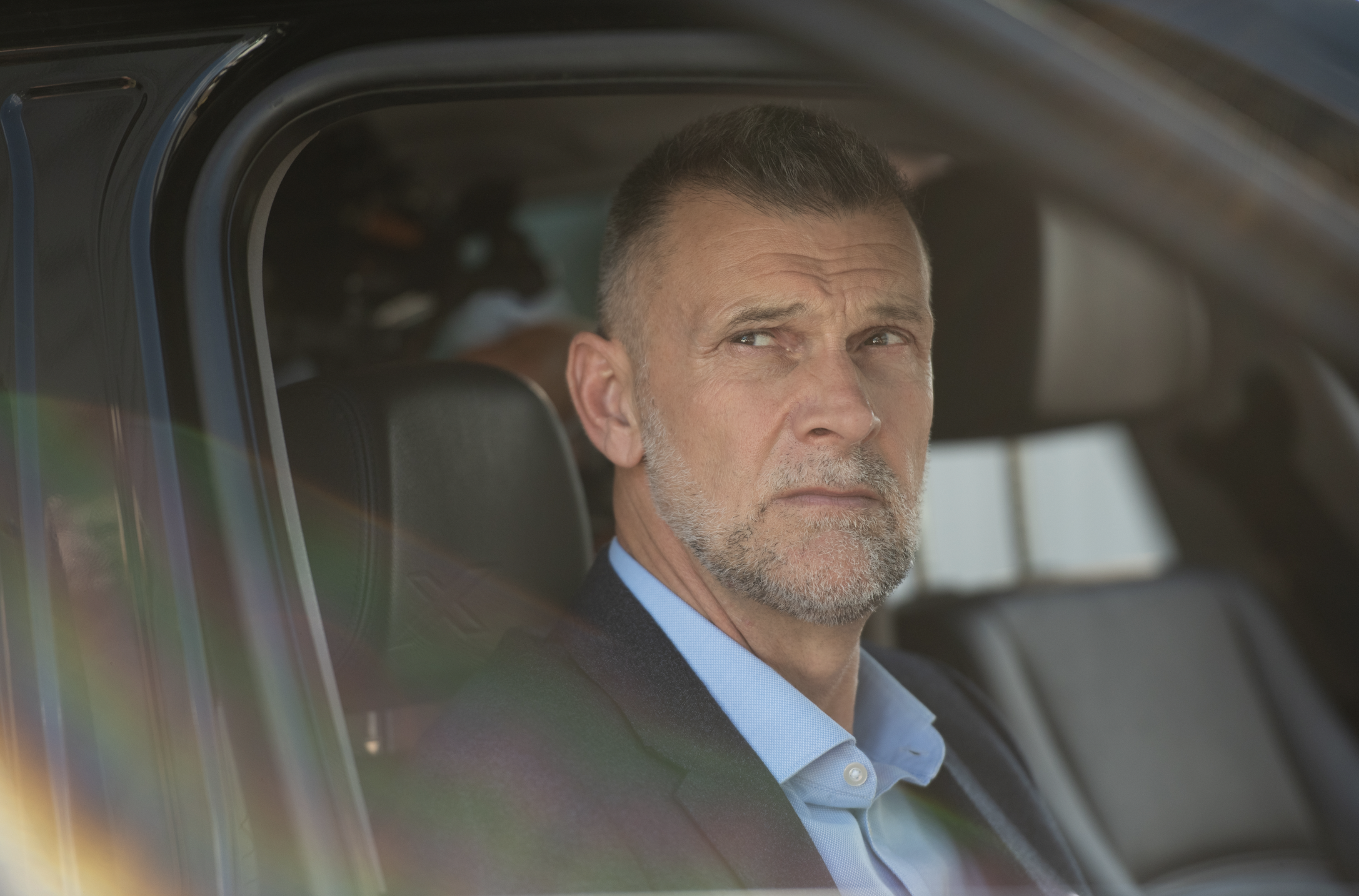 A middle-aged man with short, dark hair and a beard sitting inside a vehicle, wearing a dark suit and a light blue shirt, looking out the window with a contemplative expression.