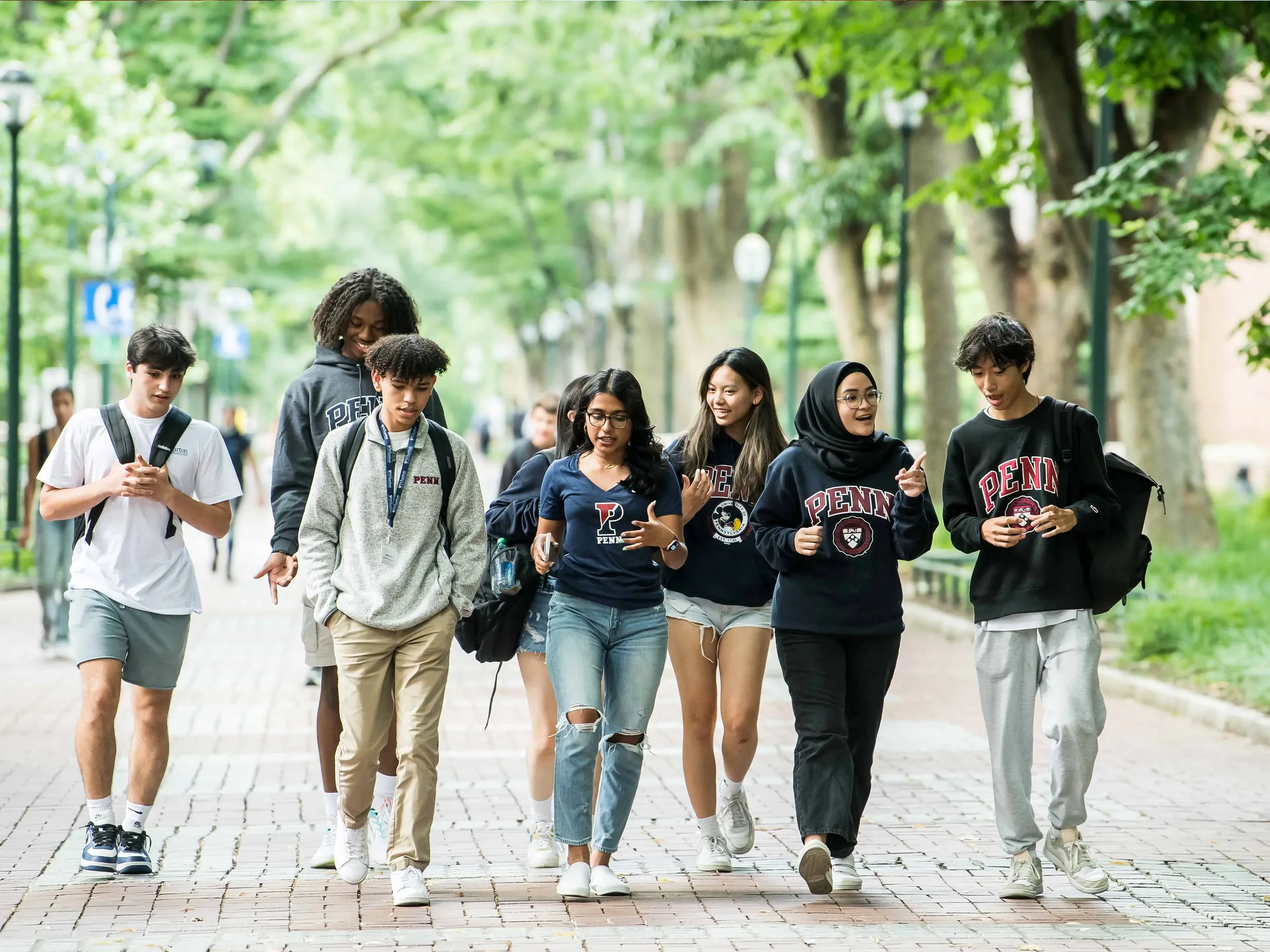 A diverse group of young people, wearing Penn and other university sweatshirts, walking and talking together on a tree-lined pathway.