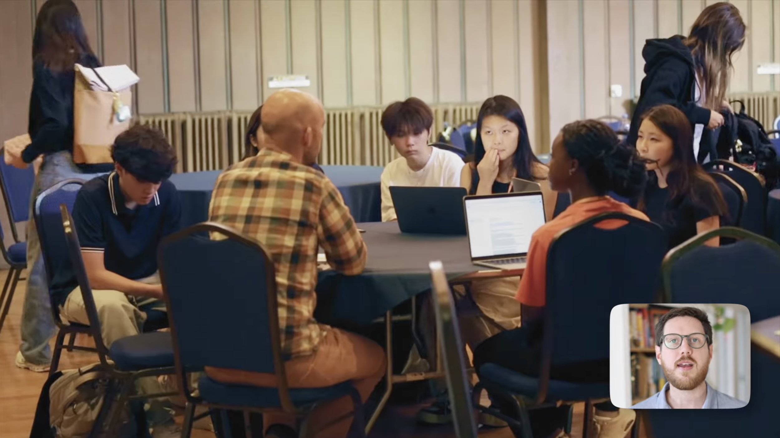 A group of young people sitting around a table in a conference room, engaging in a discussion or meeting, with two women standing at the sides.