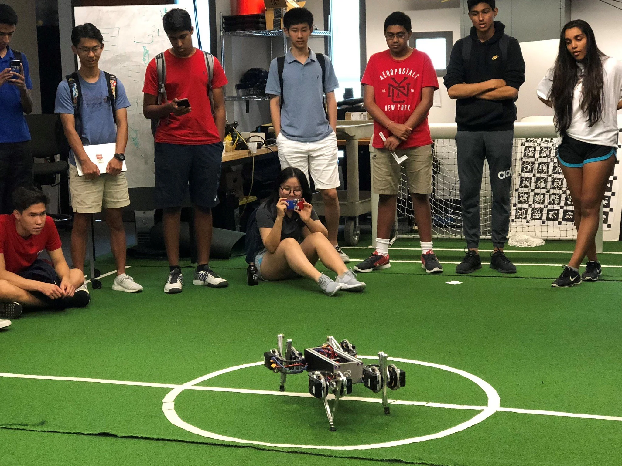 Group of teenagers at an indoor robotics workshop, observing small robots on the green turf.