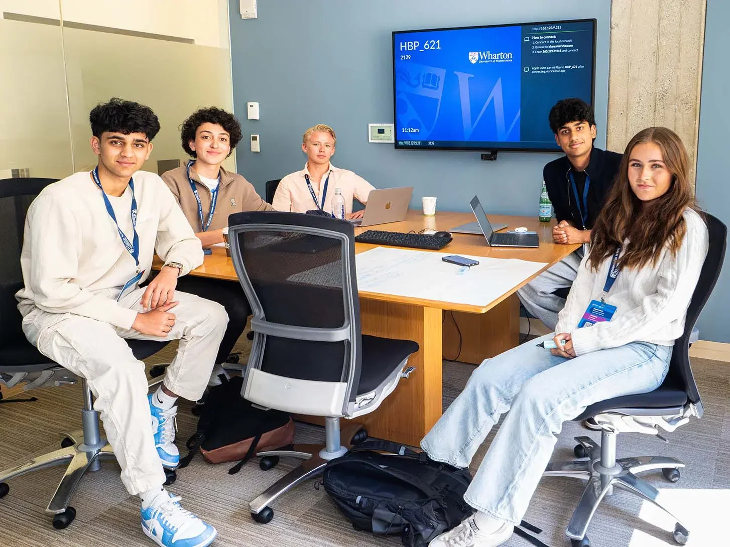 Six young students sitting around a conference table in a modern classroom, with laptops, water bottles, and notebooks, facing the camera with smiles.