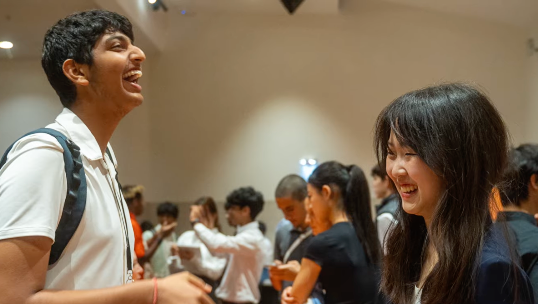 Two people smiling and laughing at a social event, with a group of others blurred in the background.