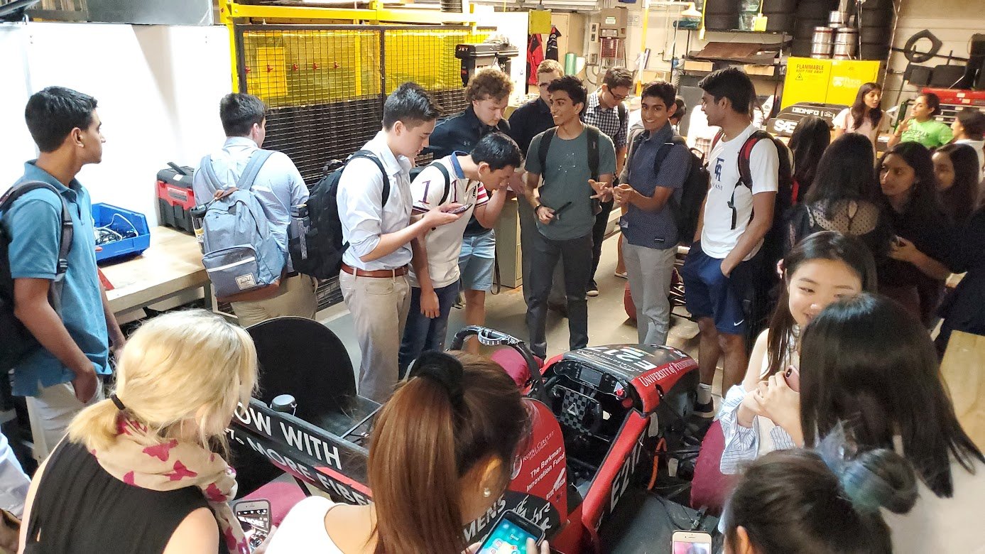 Group of students gathered around a racing car in a workshop or garage.