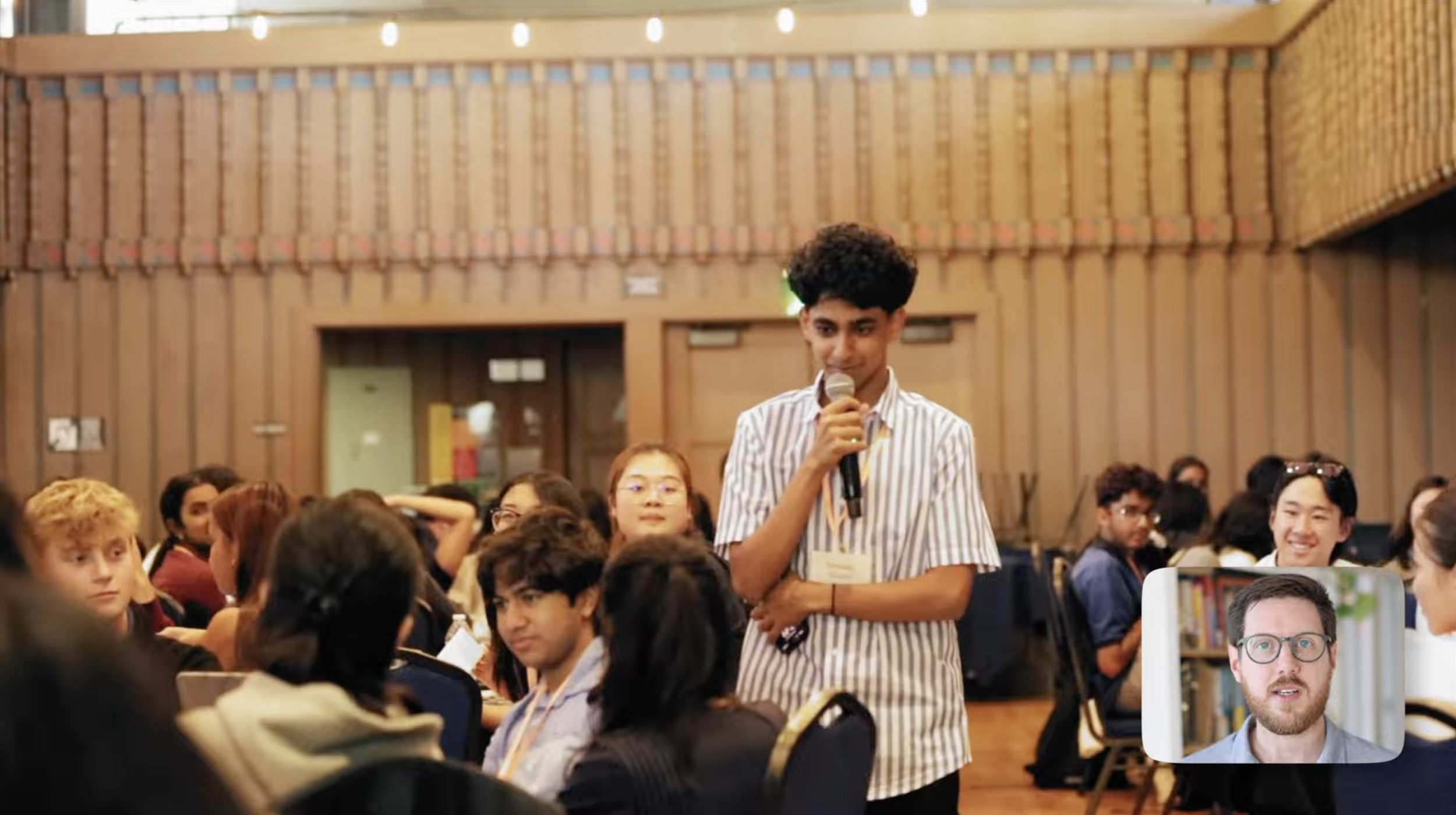 A young man with curly black hair and a striped shirt is standing among a seated audience in a large room, holding a microphone and speaking. The room has wooden walls and ceiling, with people of various ages and ethnicities listening attentively.