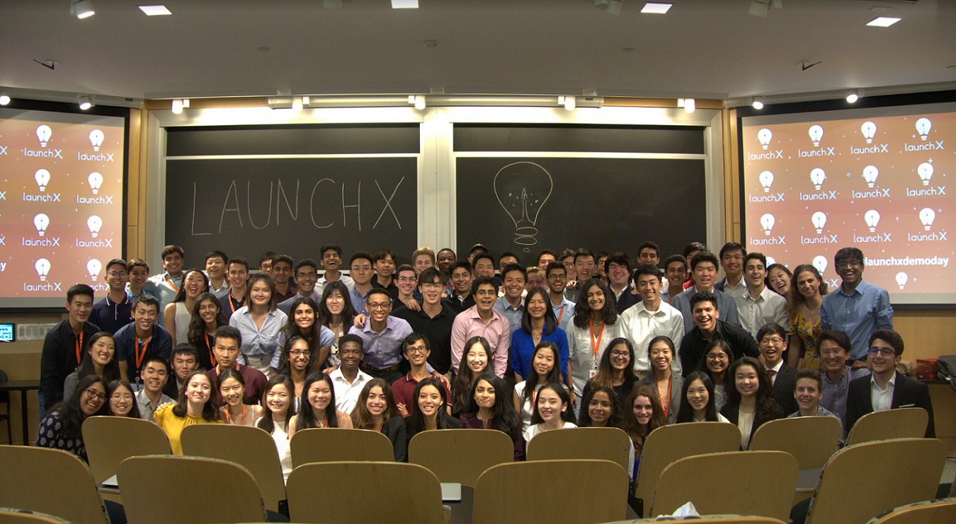 Group of students in a classroom, some wearing orange lanyards, posing for a photo in front of a blackboard that says 'LAUNCHX' with a drawing of a light bulb, and digital screens displaying 'launchX' logos.