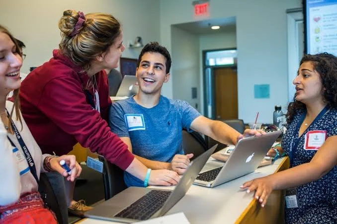 Group of young adults gathered around a table with laptops in a classroom or conference room, engaging in lively conversation and smiling.