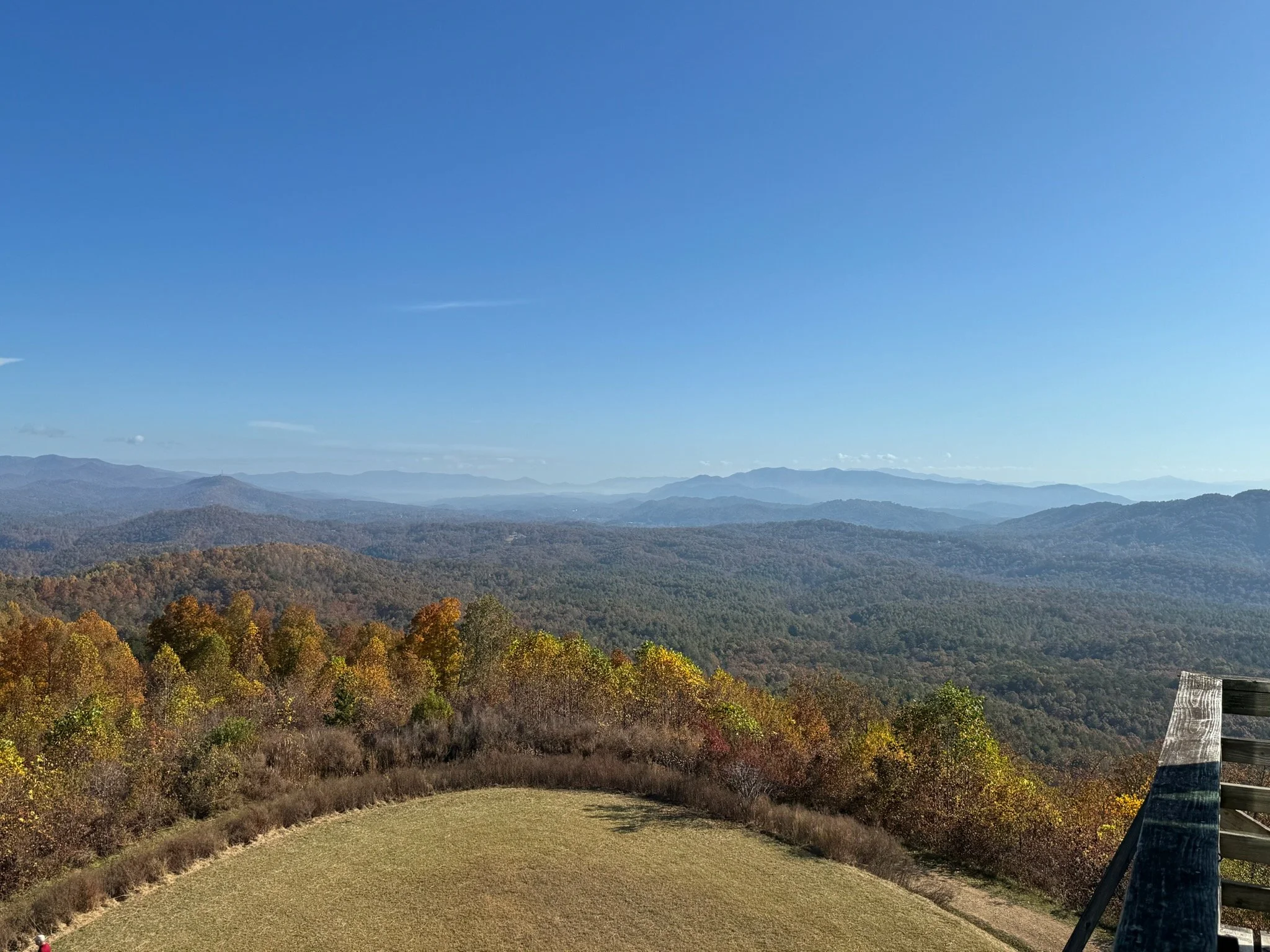 View of a mountain landscape with distant layers of mountains under a clear blue sky, with trees displaying fall foliage in the foreground and a wooden railing on the right side.