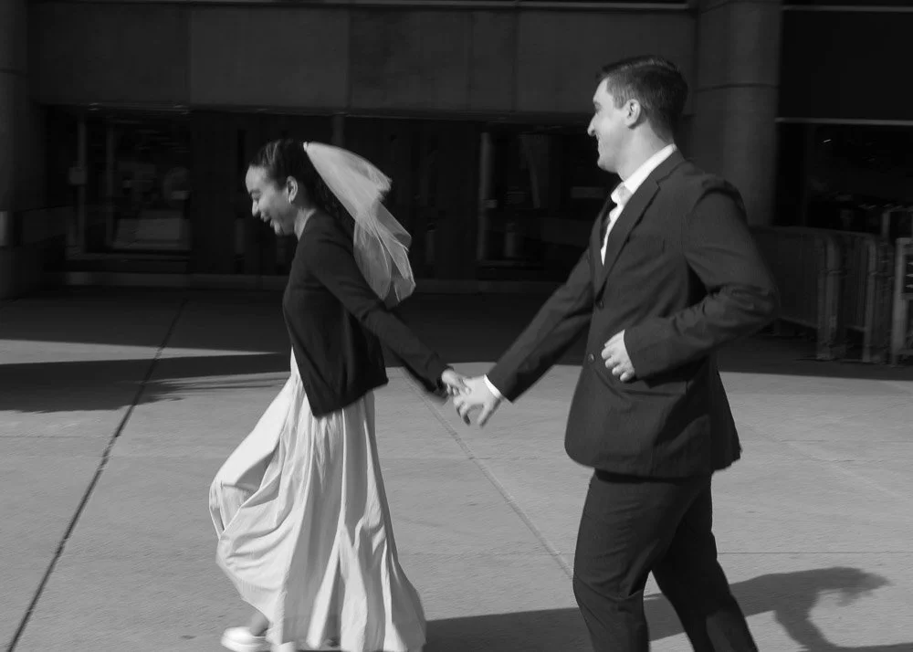 A bride and groom holding hands, walking outside, with the bride in a long wedding dress and veil, both smiling.