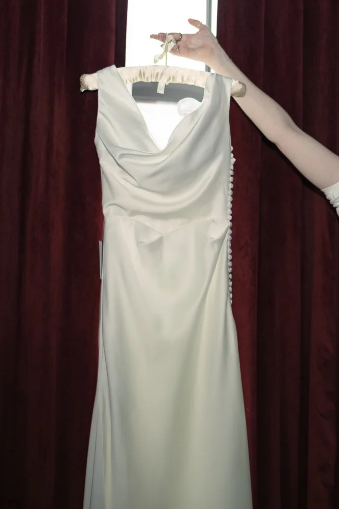 A white satin wedding dress with a Draped neckline, hanging on a hanger held by a hand, against a background of red velvet curtains.