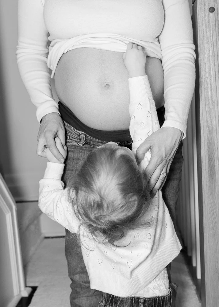 A pregnant woman standing with a young girl hugging her belly, showing an ultrasound image of the unborn baby. The woman is in a home setting.