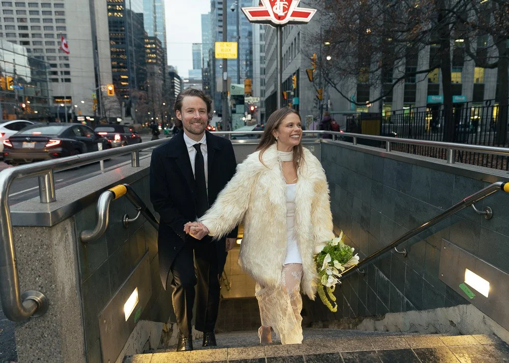 A smiling woman in a white dress with sheer lace stockings and a cream fur coat holding a bouquet of white flowers, walking up the stairs with a man in a black suit and tie, in an urban city environment with cars, buildings, and streetlights.