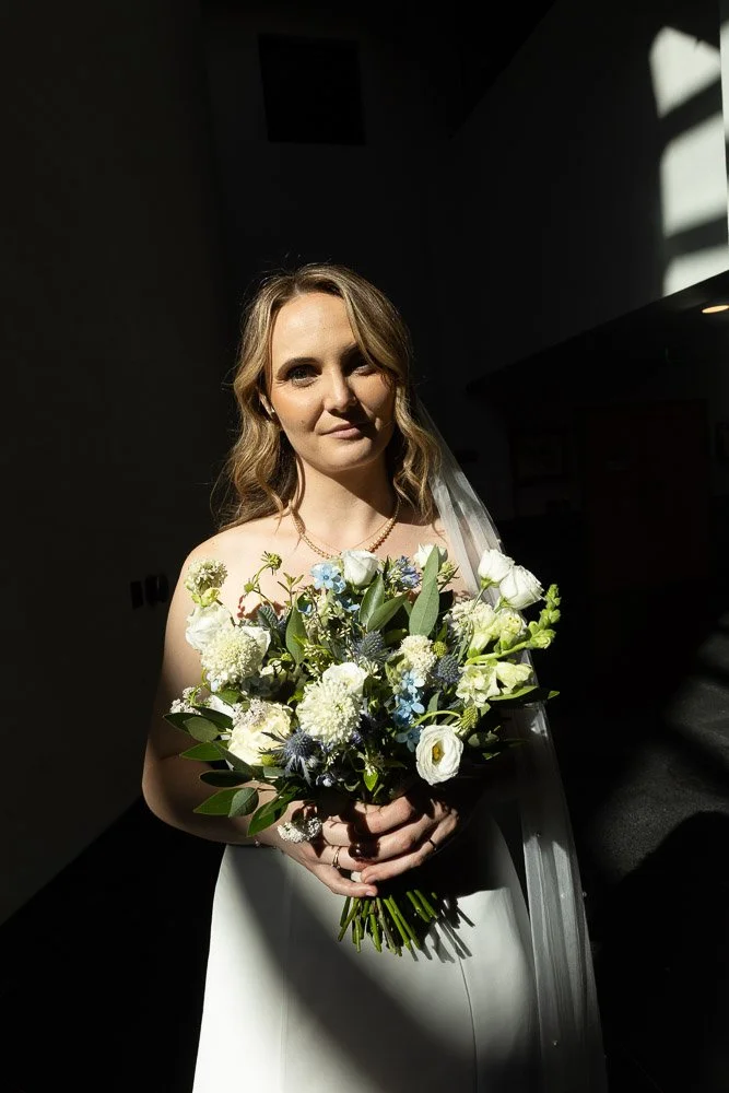 A bride with wavy blonde hair, wearing a white wedding dress, holding a bouquet of white flowers and greenery, standing in partial sunlight indoors.