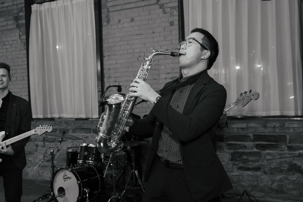 A man in glasses playing a saxophone during a band performance in a dimly lit venue with brick walls and curtains.