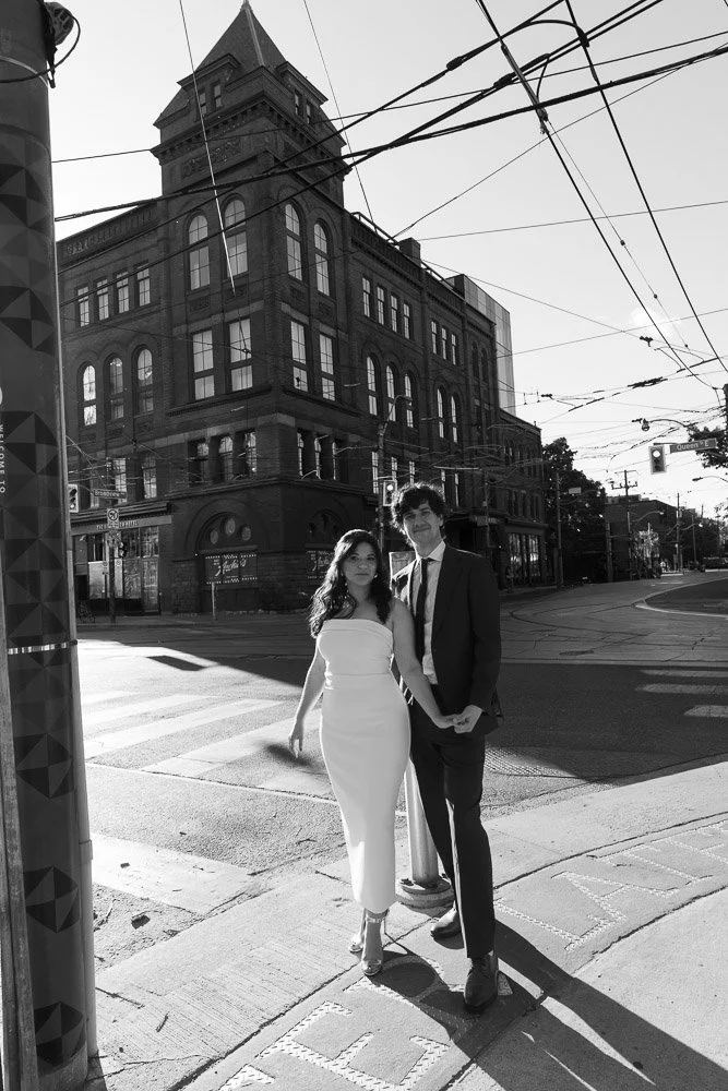A black and white photo of a couple in formal attire standing on a city sidewalk, with a historic brick building in the background and overhead streetcar wires.