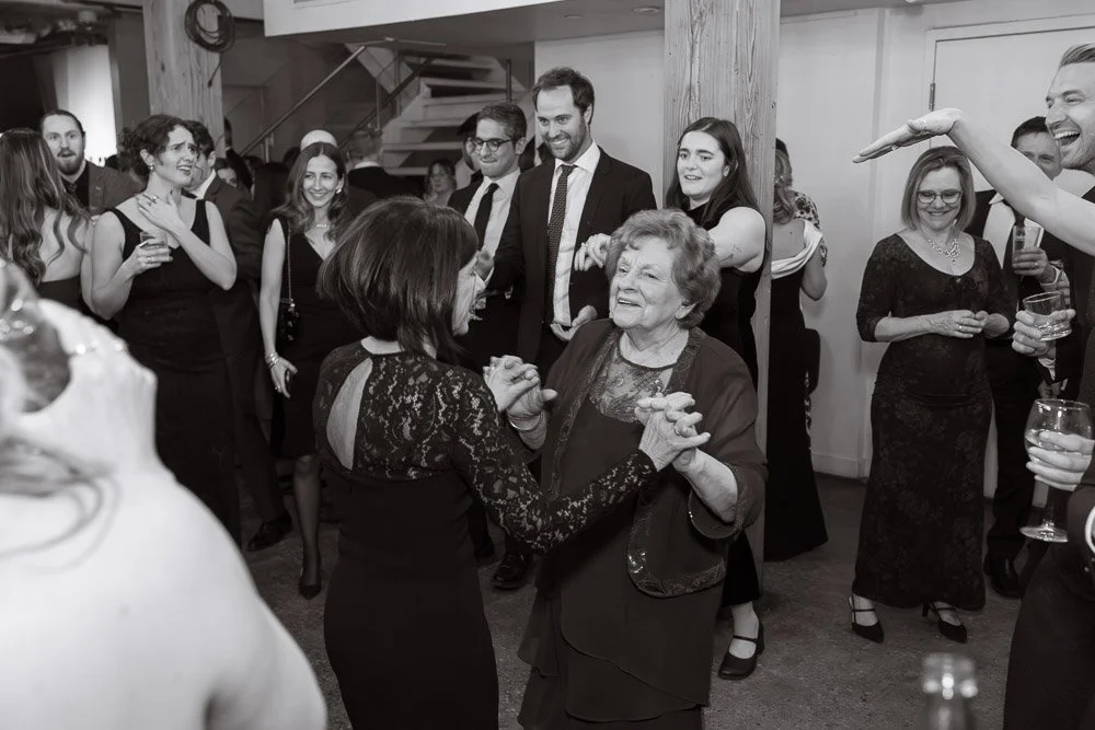 People dancing and socializing at a celebration, with an elderly woman enjoying dancing with a younger woman, while others watch and smile.