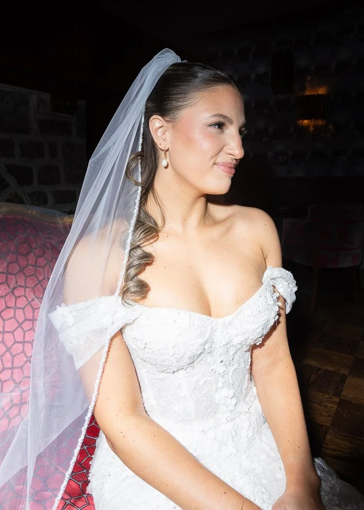 A bride with a white lace wedding dress and veil sitting indoors with a dark stone wall in the background.