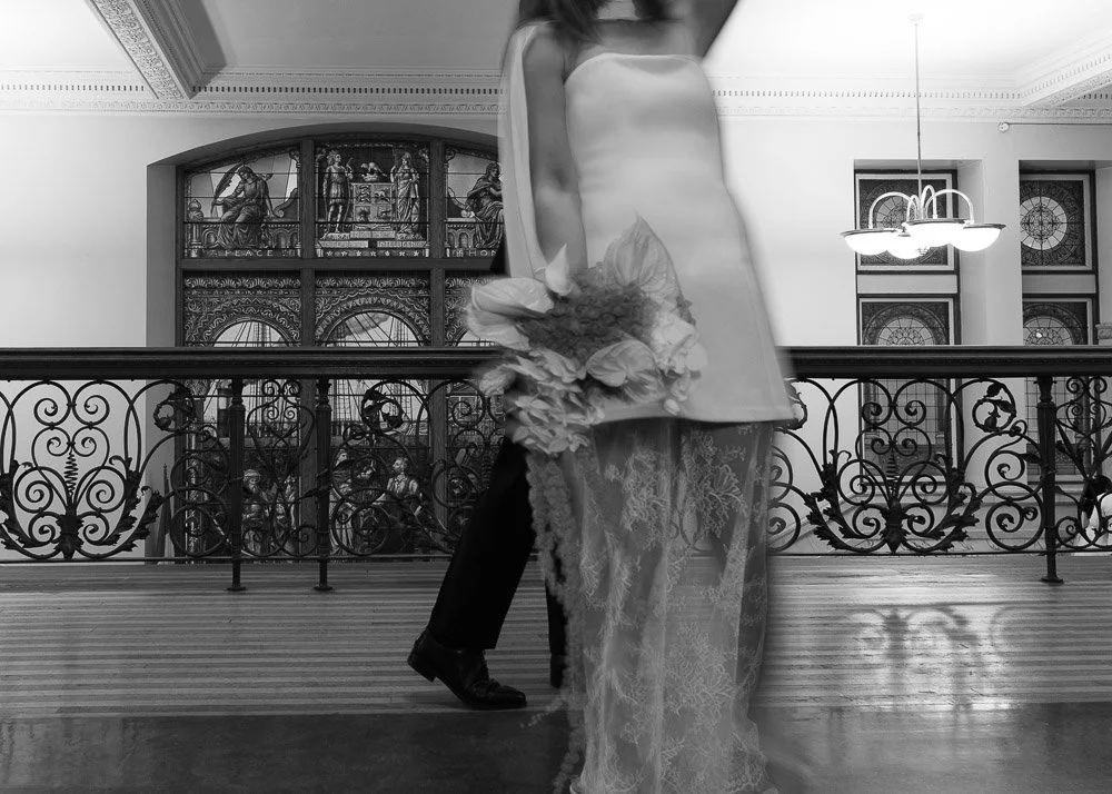 A woman in a sleeveless dress holding a bouquet of flowers standing next to a man in a tuxedo inside a historic building with ornate metal railings, stained glass windows, and a chandelier.