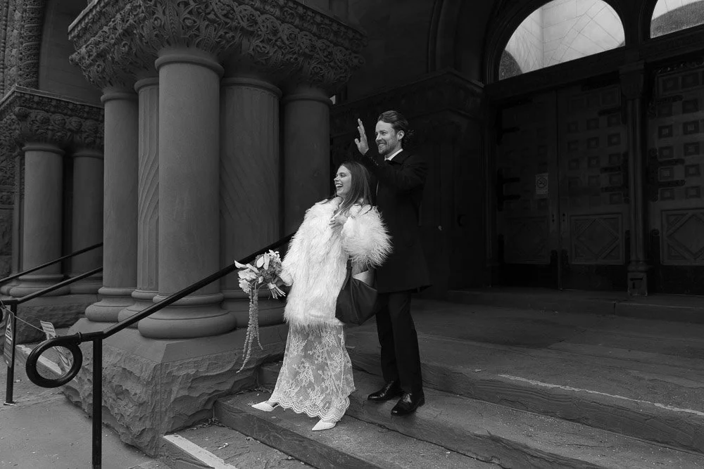 A joyful woman in a wedding dress and fur coat holding a bouquet, accompanied by a smiling man in formal attire, standing on steps outside a historic building with large columns.