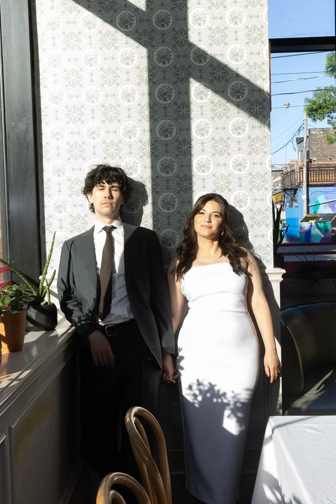 A young man in a black suit and tie standing next to a young woman in a white strapless dress, both posing indoors with sunlight casting their shadows on the wall behind them.