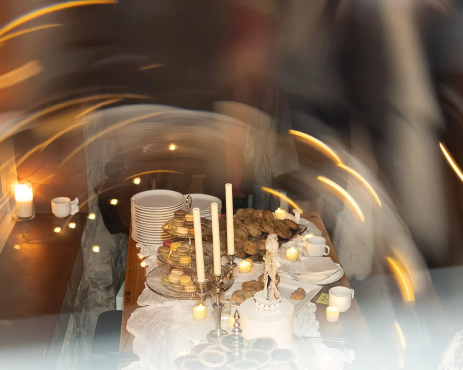 A table set with cookies, macarons, candles, and plates in a cozy indoor setting with blurred yellow light streaks.