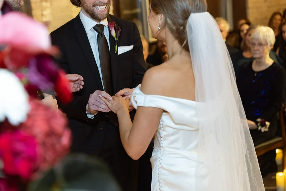 A bride and groom exchanging rings during their wedding ceremony, with guests seated in the background.