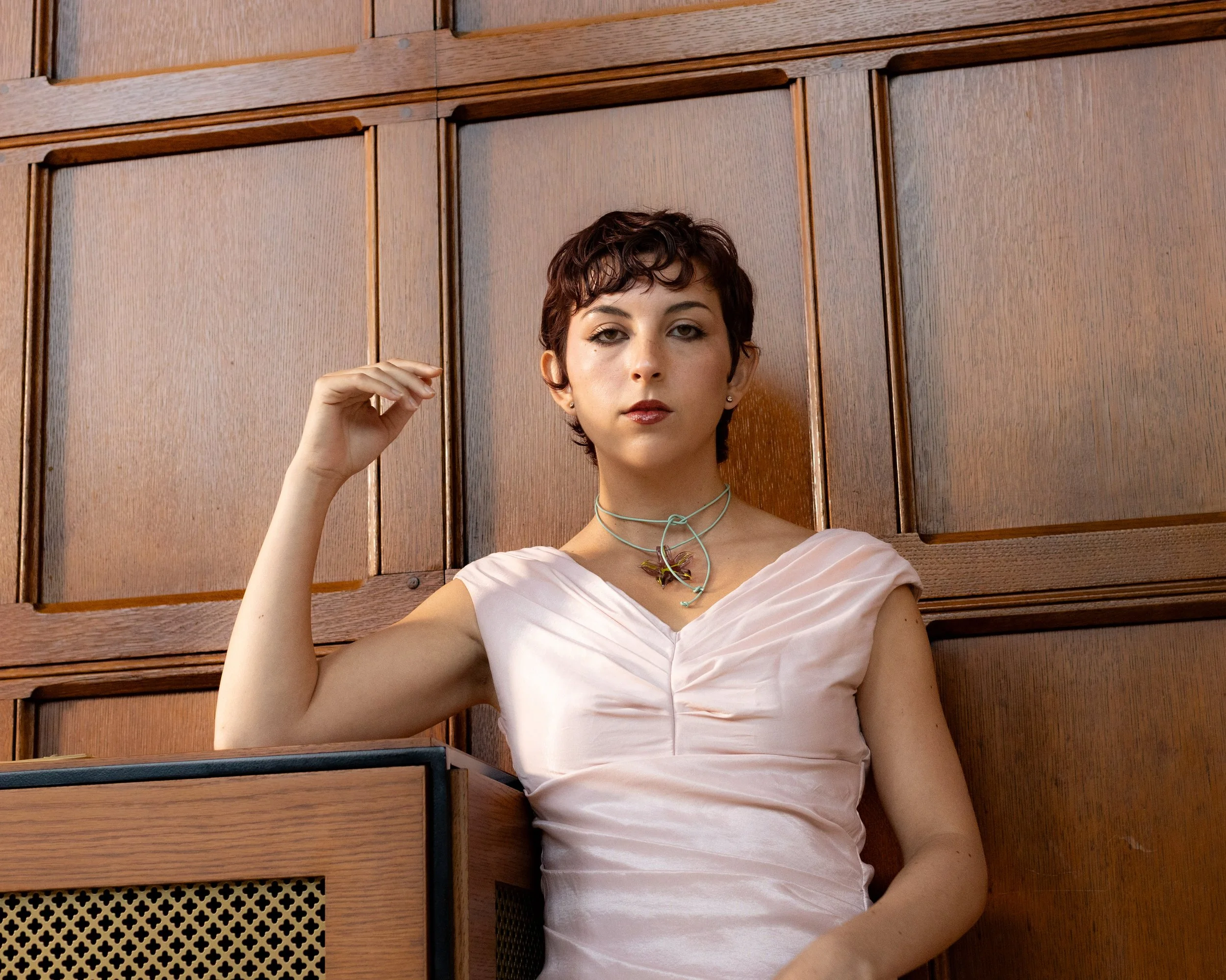 A woman with short curly hair and light skin tone sitting against a wooden paneled wall, wearing a light pink satin dress and a necklace, looking directly at the camera with a neutral expression.