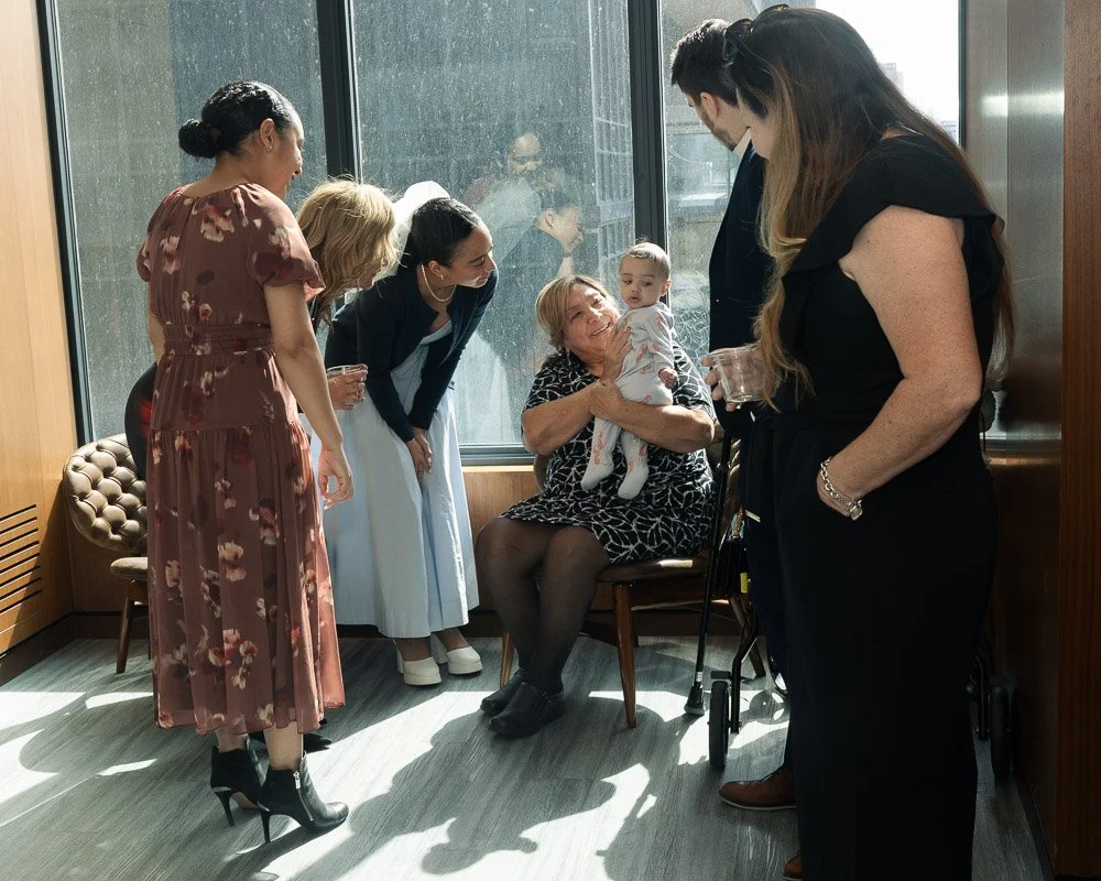 A group of women, including an elderly woman, and a baby are gathered near a large window, engaging in conversation and sharing a joyful moment, with sunlight streaming in.