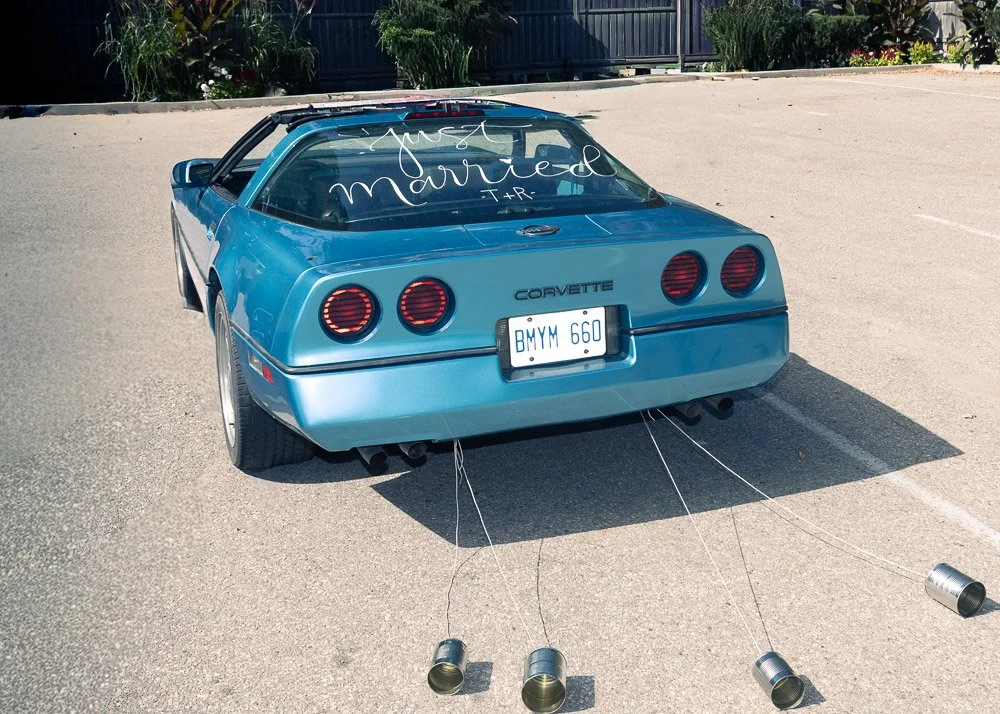 A blue Chevrolet Corvette parked outdoors on a paved surface with four cans tied to the rear bumper, connected by strings, and a handwritten message on the rear window that says 'just married'.
