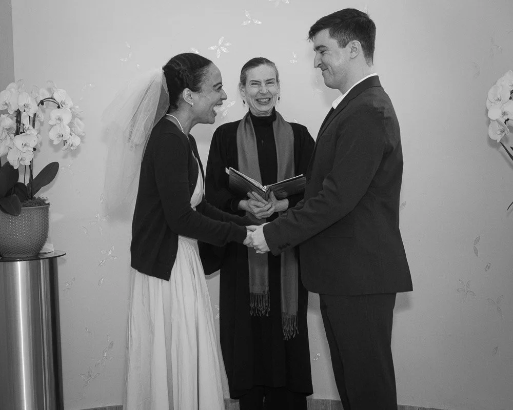 A bride and groom holding hands during their wedding ceremony, smiling at each other, with an officiant standing behind them holding a book, inside a decorated indoor venue.