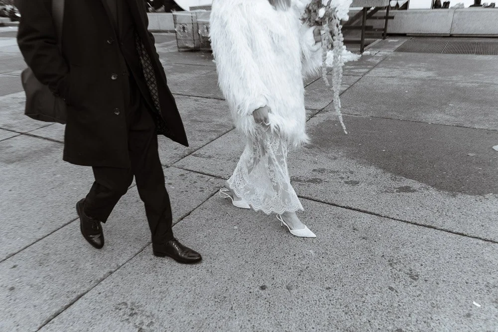 A person wearing a black coat and black shoes walking beside another person dressed in white, with only their legs visible. The person in white is wearing white high heels, a white lace dress, and a fluffy white jacket, standing on a wet sidewalk.
