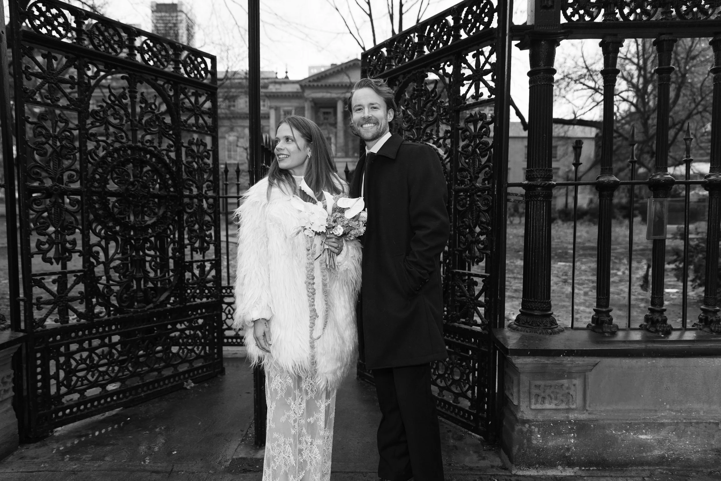 A couple dressed in formal attire standing in front of an ornate black wrought iron gate with a historic building and leafless trees in the background, on a cloudy day.