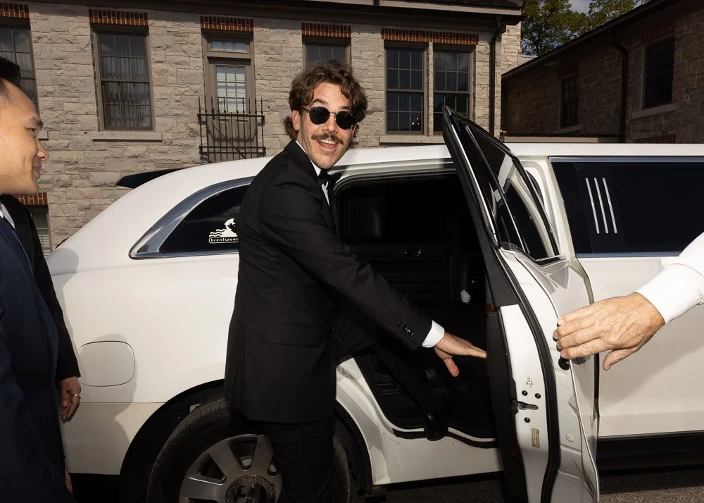 A man with curly hair, sunglasses, and a mustache wearing a black suit opening a white limousine's door, with two other men nearby, in front of a brick building.