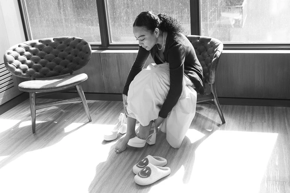 A woman sitting on a chair by a window, trying on high-heeled shoes, with a pair of slippers on the floor in front of her.