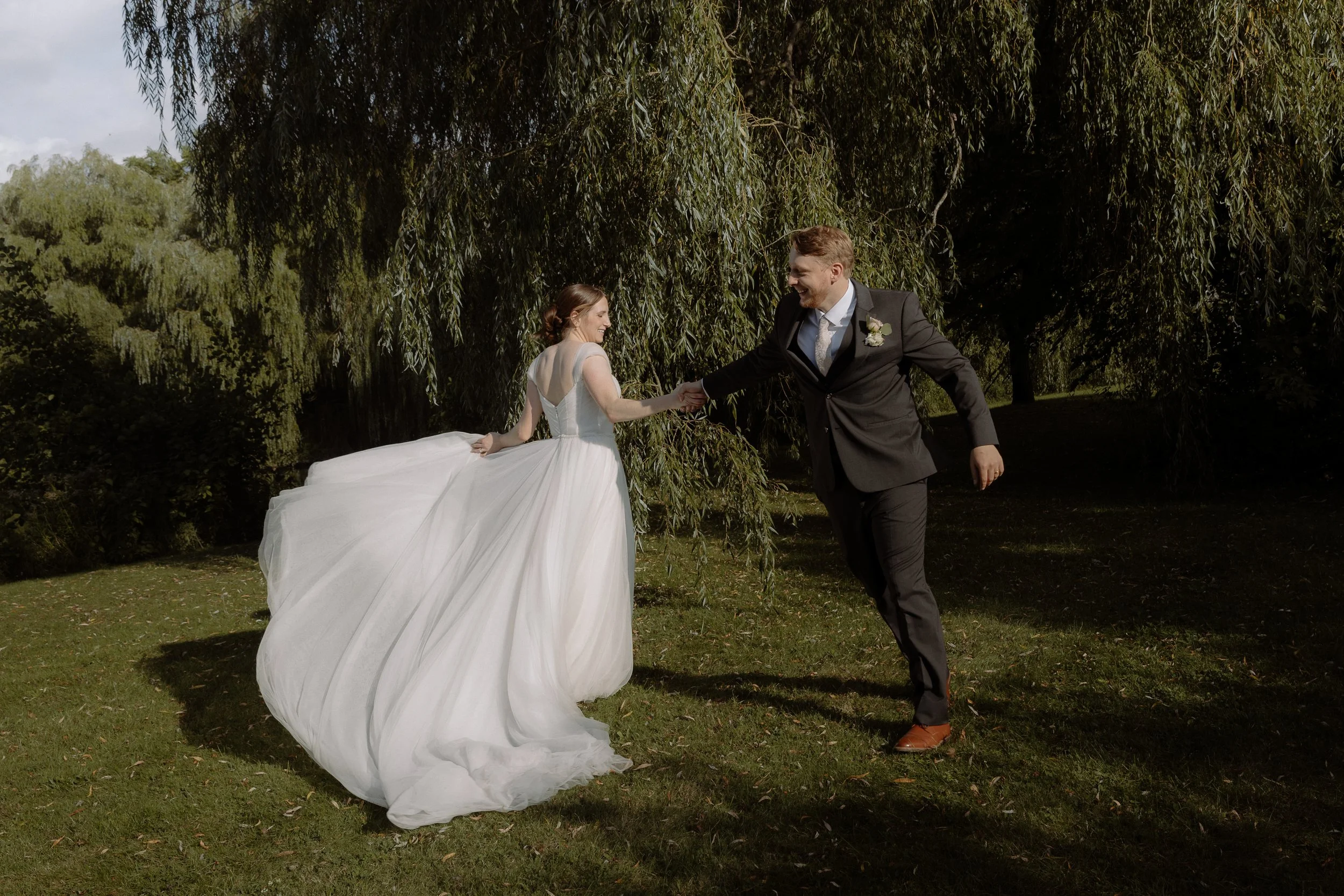 A bride and groom holding hands outdoors surrounded by trees, smiling and dressed in wedding attire.