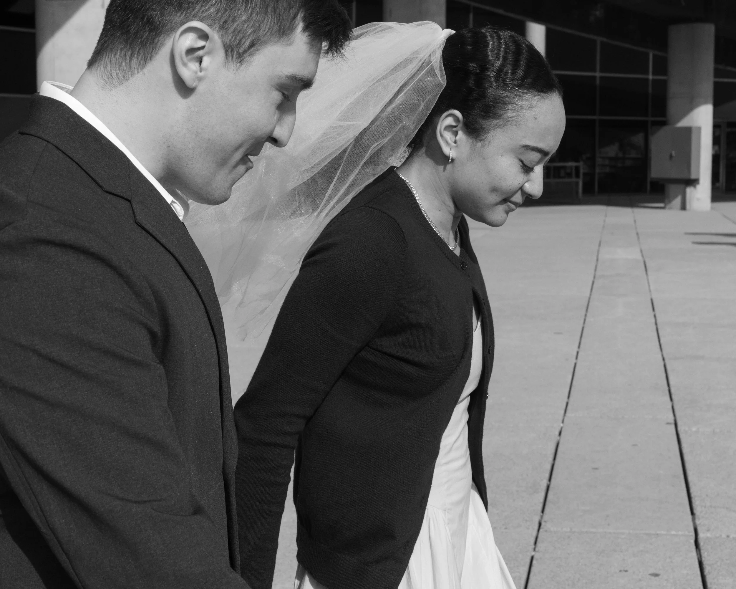 A black and white photo of a man and woman with their foreheads touching, eyes closed, with the woman wearing a veil and earrings, and the man in a suit. They appear to be engaged in a moment of intimacy or prayer outside a building.