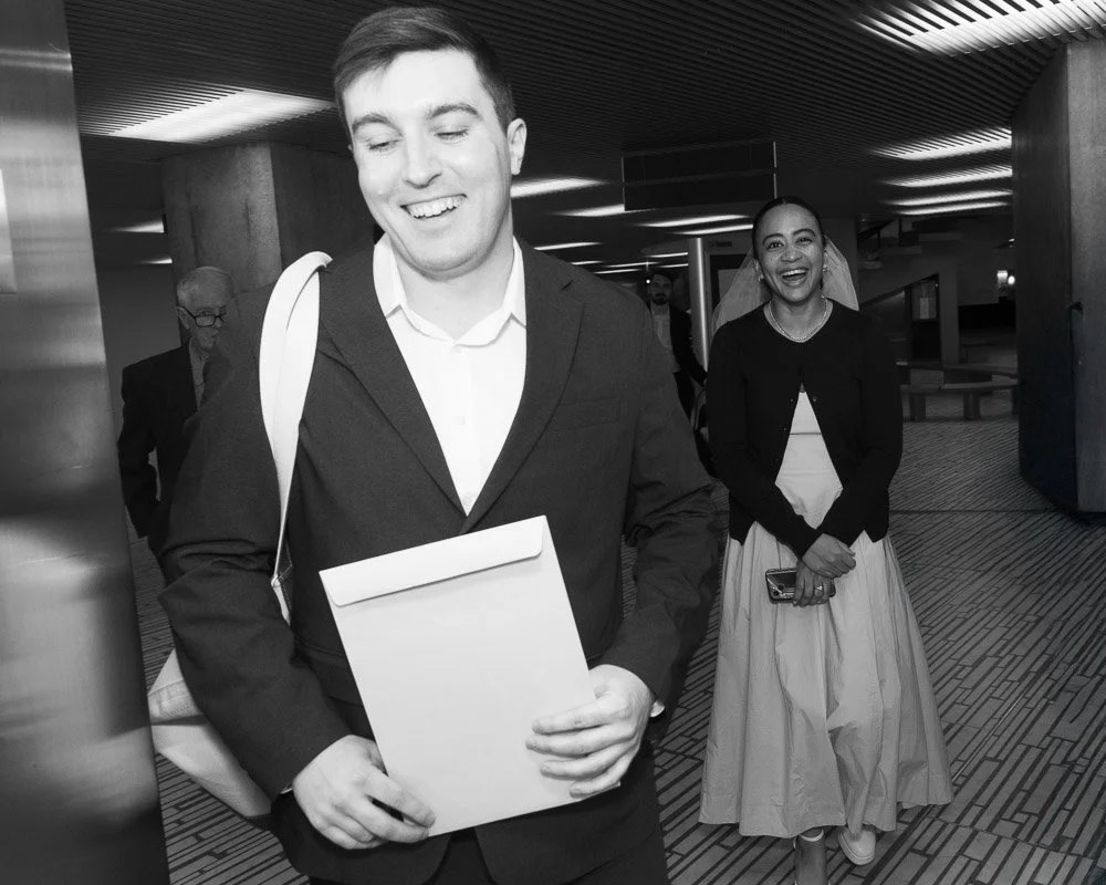 A happy man in a suit carrying an envelope and a bag, walking in an indoor space with other people, including a woman smiling and holding a phone, likely at an event or airport.