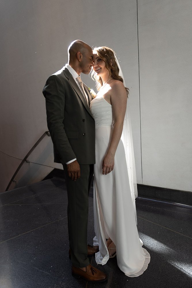 A bride and groom are standing close together, smiling at each other, inside a modern, minimalistic building. The bride is wearing a white strapless wedding dress with a veil, and the groom is in a dark suit with a light-colored shirt and tie. They are touching foreheads in an intimate moment.