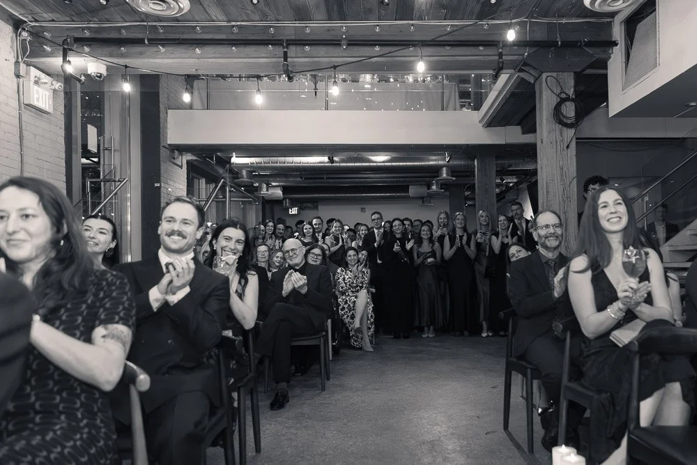 A group of people dressed in formal attire, gathered in a modern event space, watching a performance or presentation, with some seated and others standing and clapping.