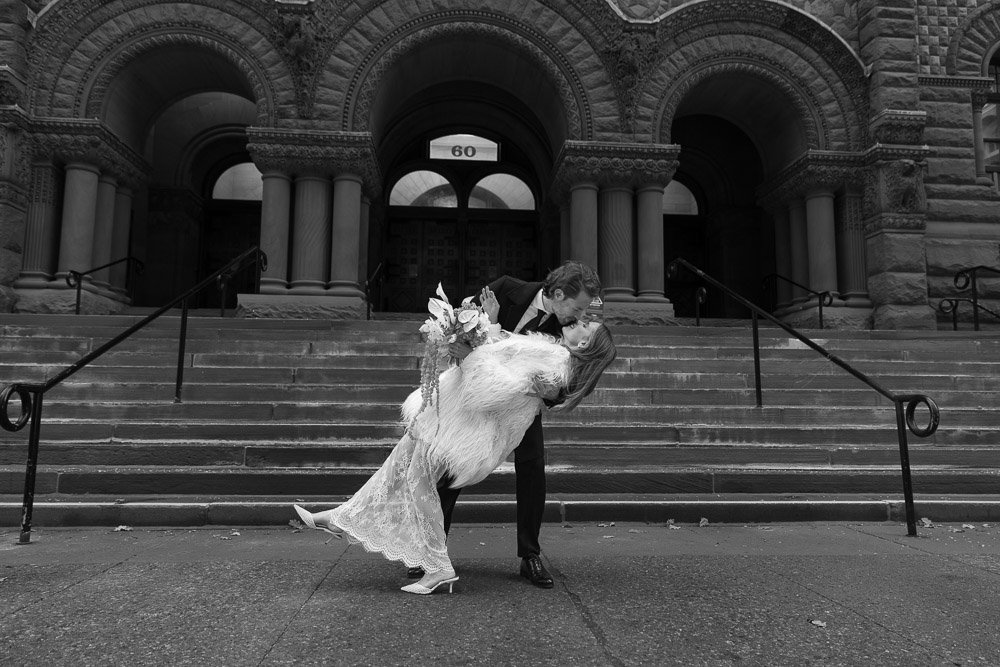A couple sharing a kiss on the steps of a historic stone building, with the man holding the woman in a dip. The woman is in a wedding dress holding a bouquet, and the scene is in black and white.