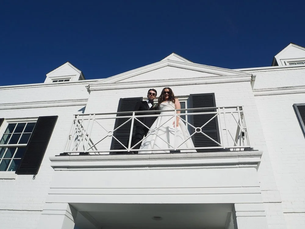 A couple dressed in formal wedding attire standing on a white balcony of a white building with black shutters, under a clear blue sky.