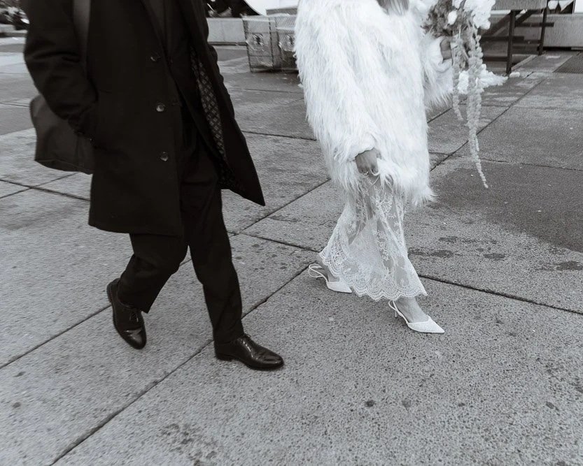 A person in a dark suit and coat, walking on a wet sidewalk, and a woman in a white fur coat and lace dress, with high heels, standing on the sidewalk.