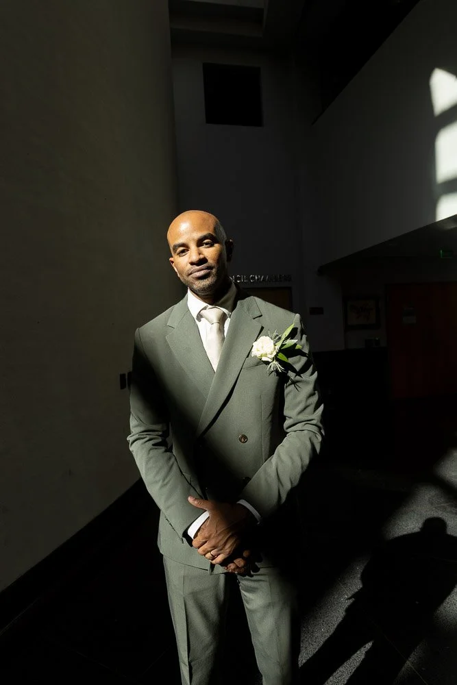 A man in a gray suit with a white shirt and boutonniere on his lapel, standing in a dimly lit indoor space with a sunlight patch casting shadows.